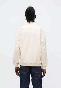 Lys beige bomberjakke med mønstret tekstur, elastiske mansjetter og kant, samt en liten etikett på baksiden. Kombinert med mørkeblå jeans.