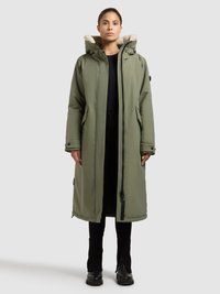 Lange olijfgroene jas met een met bont gevoerde capuchon, ritssluiting, zijzakken en zwarte accenten, gedragen over een zwarte outfit en enkellaarzen.