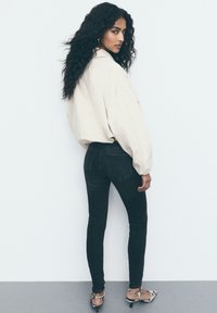 Beige oversized shirt, zwarte high-waisted jeans en gemêleerde haksandalen. Krullend haar los gestyled; model kijkt iets achterover.