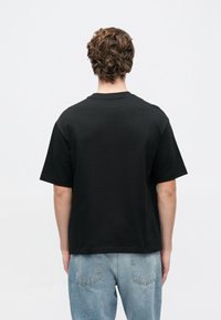 Čierne bavlnené tričko s krátkymi rukávmi, okrúhlym výstrihom a voľným strihom, zobrazené zozadu, v kombinácii so svetlomodrými džínsami.