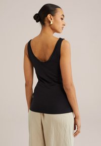 Czarna koszulka na ramiączkach z niskim dekoltem w kształcie litery V i koronkowym wykończeniem, wykonana z miękkiego materiału. Modelka pokazana jest od tyłu, nosząc jasne spodnie.