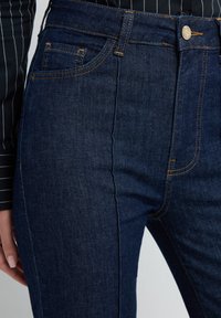 Tmavě modré džínové kalhoty s úzkým střihem, zlatým prošitím, přední kapsou a kovovým zapínáním na knoflík v pase.