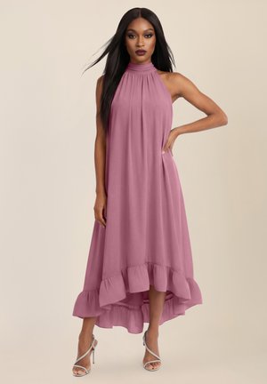 Vrouw met lang zwart haar, gekleed in een mouwloze mauve jurk met hoge hals, ruches aan de zoom en zilveren sandalen met bandjes, hand op de heup.