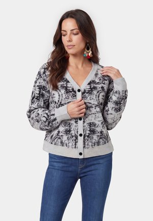 Kvinde iført en grå og sort mønstret knap-op cardigan med ribbet manchetter, kombineret med blå jeans og farverige frynseøreringe.