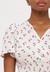Hvit omslagsbluse med rødt kirsebærblomstermønster, korte puff-ermer og smocket midje. Lett stoff med en myk, teksturert finish.
