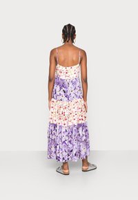 Femme avec des tresses portant des boucles d'oreilles créoles et une robe florale à volants en violet et blanc, se tenant face à un fond léger uni.