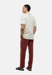 Polo shirt beige clair à manches courtes, associé à un pantalon couleur rouille. Le modèle se tient avec les mains dans les poches, portant des sandales noires.