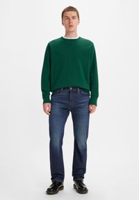 Grüner Pullover mit Rundhalsausschnitt, kombiniert mit dunkelblauen Jeans und schwarzem Schuhwerk, vor einem schlichten weißen Hintergrund stehend.