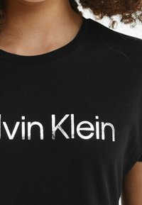 Nahaufnahme einer Person, die ein schwarzes T-Shirt mit dem weißen Calvin Klein-Logo auf der Brust trägt.