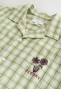 Chemise à manches courtes en tartan vert clair avec boutons, col, fleurs violettes brodées et texte "FLEURS" sur la poitrine gauche.