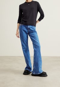 Suéter de punto negro con cuello redondo, combinado con unos jeans acampanados azules y unos zapatos negros de lona texturizada sin cordones. Diseño sencillo, corte clásico.