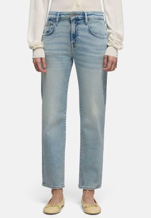 Person trägt hellblaue Jeans mit geradem Bein, off-white Langarmshirt und blassgelbe Ballettschuhe, steht vor einem einfarbigen Hintergrund.