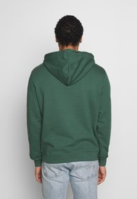 Felpa verde scuro con cappuccio regolabile, maniche lunghe e polsini e orlo a coste. Tessuto liscio. Indossata con jeans blu chiaro.
