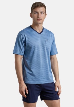 Young man wearing a blue patterned V-neck short-sleeve shirt with a chest pocket and dark blue shorts standing against a plain background.