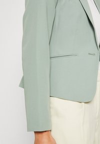 Světle zelená přizpůsobená blazer s hladkou texturou, jednobuším zapínáním a křivým lemem. Stylizováno přes krémové kalhoty.
