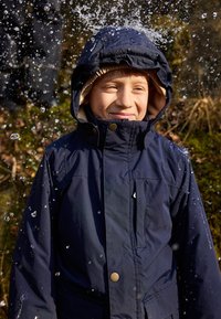 Giacca impermeabile blu navy con cappuccio bordato in marrone, bottoni visibili e tasca frontale. Gocce d'acqua schizzano intorno a chi la indossa.