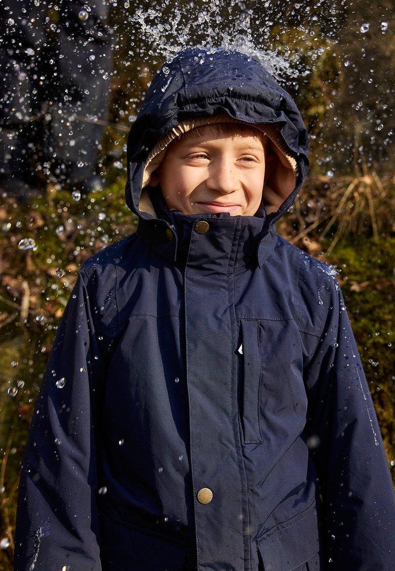 Giacca impermeabile blu navy con cappuccio bordato in marrone, bottoni visibili e tasca frontale. Gocce d'acqua schizzano intorno a chi la indossa.