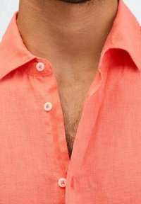 Camicia di lino corallo con colletto a punta, due bottoni bianchi sul collo e una finitura testurizzata. Tessuto morbido e leggero visibile.