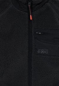Black fleece jacket with a zippered front, textured surface, and a chest pocket. Features an orange zipper pull and branding detail.