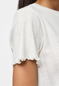 Vit texturerad topp med puffärmar, med ett fint öppet mönster och volangkant. Slät tyg med horisontella ränder.