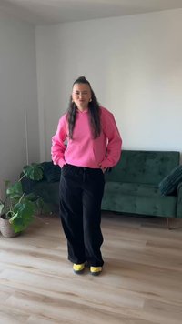 Pink hooded sweatshirt, black loose-fitting pants, and yellow shoes on a person standing in a room with a green couch and plants.
