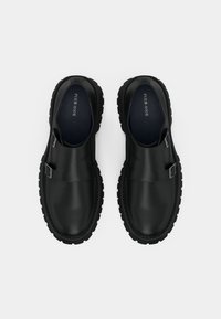 Μαύρα δερμάτινα παπούτσια τύπου slip-on με χοντρή καουτσούκ σόλα, διαθέτουν δύο ρυθμιζόμενες ασημένιες αγκράφες και λείο, γυαλιστερό φινίρισμα.