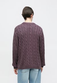 Μωβ πλεκτό πουλόβερ με σχέδιο καλωδίου, με ριμπ λαιμόκοψη και μανσέτες, σε άνετη γραμμή, φορεμένο πάνω από ανοιχτόχρωμο μπλε τζιν.