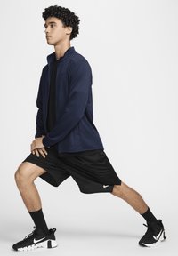 Chaqueta atlética de color navy, camiseta negra, pantalones cortos negros y zapatillas negras con detalles en blanco. Modelo mostrado en una pose de estocada contra un fondo blanco.