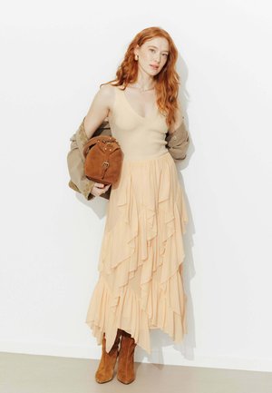 Femme aux cheveux roux portant un haut beige sans manches, une jupe pêche superposée, des bottes en daim beige, tenant un sac en daim marron et une veste kaki posée sur son bras.