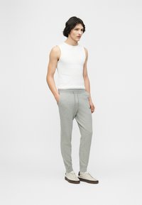 Άνδρας με κοντά σκούρα μαλλιά, ντυμένος με λευκή κοντομάνικη μπλούζα, ανοιχτογκρίζα παντελόνια φόρμας με τσέπες και μπεζ αθλητικά παπούτσια, στέκεται με τα χέρια στις τσέπες.