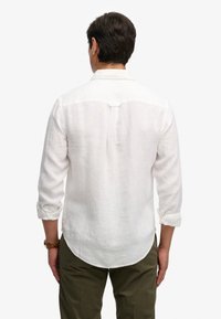 Hombre de espaldas vestido con una camisa blanca de lino de manga larga con los puños remangados y pantalones verde oliva, sobre un fondo liso.