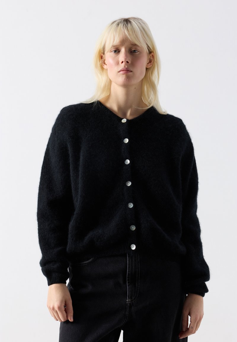 Blondína oblečená v čiernom huňatom kardigane na gombíky a čiernych nohaviciach, stojaca pred jednoduchým svetlosivým pozadím.