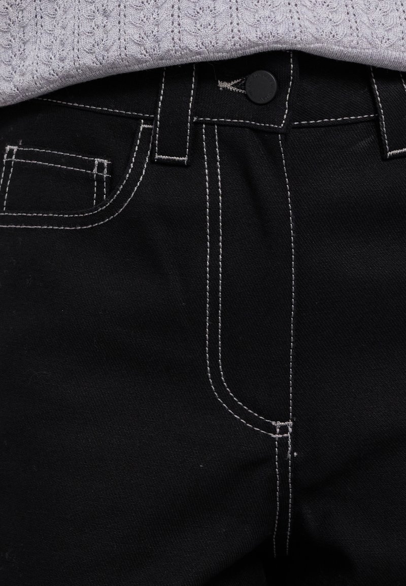 Pantaloni in denim neri con cuciture bianche a contrasto, dotati di chiusura con bottone, passanti per cintura e una tasca frontale sul lato destro.