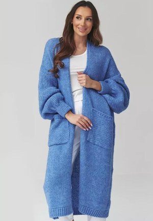 Kobieta w długim, grubym niebieskim swetrze na drutach, nałożonym na biały top i białe spodnie, stojąca na tle jednolitego szarego tła.