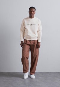Crème sweatshirt met logo, gecombineerd met losse bruine broeken en witte sneakers. Gladde textuur, relaxte fit, minimalistisch ontwerp.