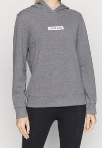 Persoon in een grijze Calvin Klein hoodie en zwarte legging met subtiele tekst op de dij, staande tegen een effen achtergrond.