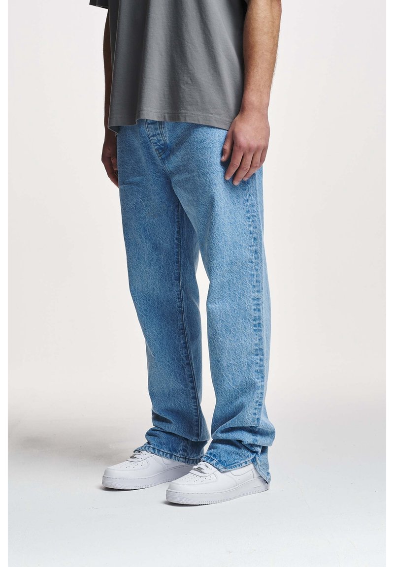 Persona vestida con jeans de corte amplio de color azul claro, camiseta gris y zapatillas blancas, de pie sobre un fondo blanco liso.