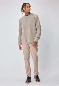 Homme aux dreadlocks portant un pull beige, un pantalon kaki clair et des chaussures noires à lacets, debout devant un fond clair uni.