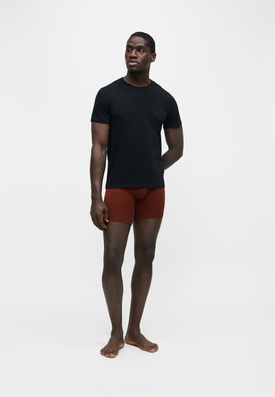 Homme debout pieds nus portant un t-shirt noir moulant à manches courtes et un short moulant couleur rouille sur un fond clair uni.