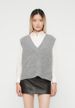 Jonge vrouw in een witte blouse, grijze gebreide mouwloze vest, en een korte zwarte leren rok, staand tegen een effen achtergrond.