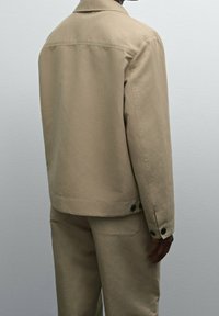 Giacca in cotone beige con colletto strutturato, spalla posteriore, dettagli con bottoni, taglio dritto e design minimalista. Vestibilità su misura.