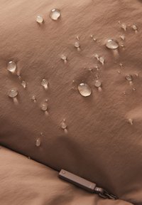 Braune Stoffoberfläche mit Wassertropfen, die eine glatte Textur zeigt. Ein kleiner brauner Reißverschlusszipp ist teilweise in der unteren rechten Ecke sichtbar.