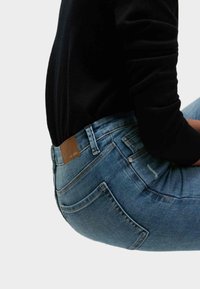 Suéter oscuro combinado con jeans de mezclilla ajustados de color azul claro. Los jeans tienen un parche de cuero marrón y bolsillos traseros con detalles de costura.