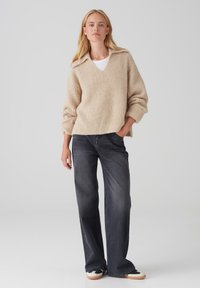 Maglione beige lavorato a maglia con scollatura a V e trama a coste, abbinato a jeans larghi grigio scuro e sneakers bianco e nero.