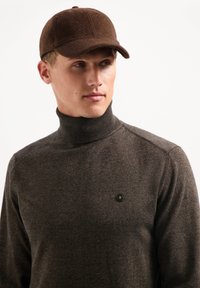 Een bruine wollen muts met een afgeronde piek, gecombineerd met een donkerbruine turtleneck trui met een klein hexagonaal logo op de linkerkant van de borst.