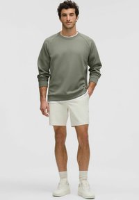 Olijfgroen sweatshirt met raglanmouwen, gecombineerd met witte shorts. Witte sneakers en lichte sokken maken de outfit compleet. Eenvoudig ontwerp, zachte textuur.