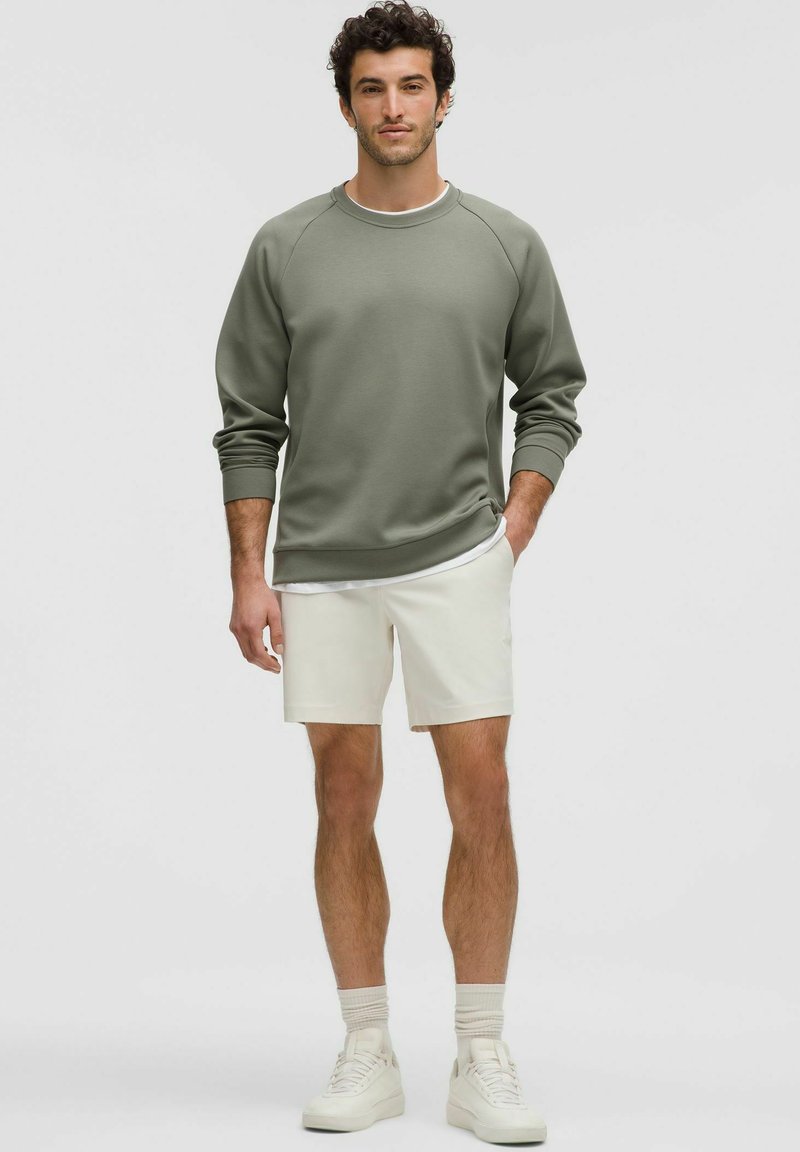 Olijfgroen sweatshirt met raglanmouwen, gecombineerd met witte shorts. Witte sneakers en lichte sokken maken de outfit compleet. Eenvoudig ontwerp, zachte textuur.