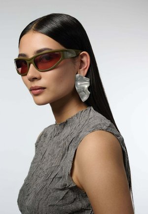 Femme aux longs cheveux foncés portant des lunettes de soleil angulaires vert olive, de grandes boucles d'oreilles argentées abstraites et un haut sans manches gris texturé.