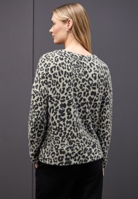 Nainen, jolla on pitkät vaaleat hiukset, pukeutuneena pörröiseen leopardikuvioiseen neuleeseen ja mustiin housuihin, seisoo sivuttain yksinkertaista tummanharmaata taustaa vasten.