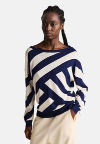 Námořnický a krémově pruhovaný svetr s širokým výstřihem, měkkou strukturou a volným střihem, v kombinaci se světlými kalhotami.